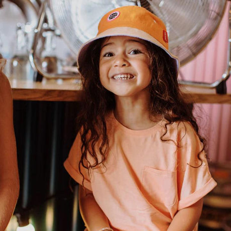 Lachend kind met het oranje hoedje Mini Orange van Hello Hossy en een perzikkleurig T-shirt.