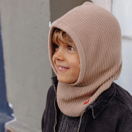 Kind met beige gebreide Hoody Latte van Hello Hossy over het hoofd en rond de hals, boven een donkere jas.