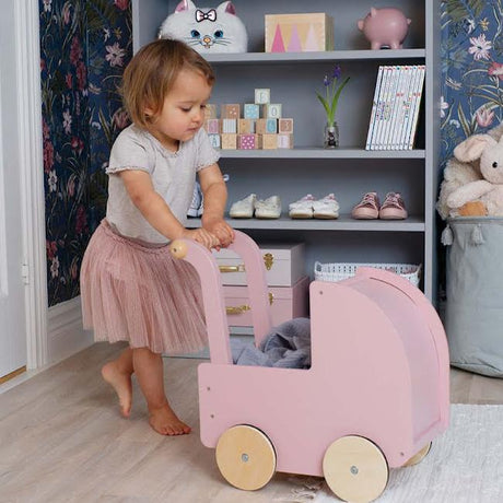 Kind duwt een roze houten poppenwagen met naturel houten wielen in een kinderkamer.