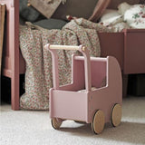 Roze houten poppenwagen met naturel houten wielen op een vloerkleed naast een bed met een bloemendeken.