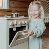 Kind opent de ovendeur van de beige houten speelkeuken met een pannetje op het fornuis.