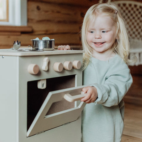 Kind opent de ovendeur van de beige houten speelkeuken met een pannetje op het fornuis.