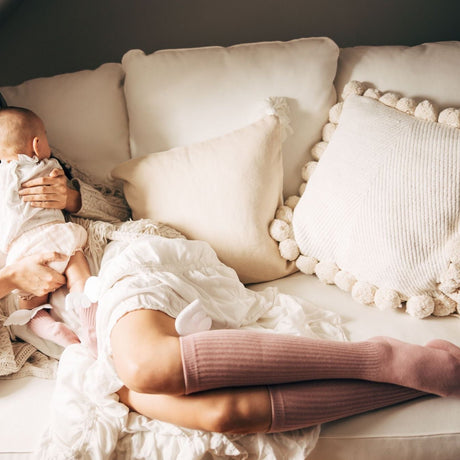 Mama draagt roze kniekousen met witte vleugeltjes en houdt een baby vast op een lichte bank.
