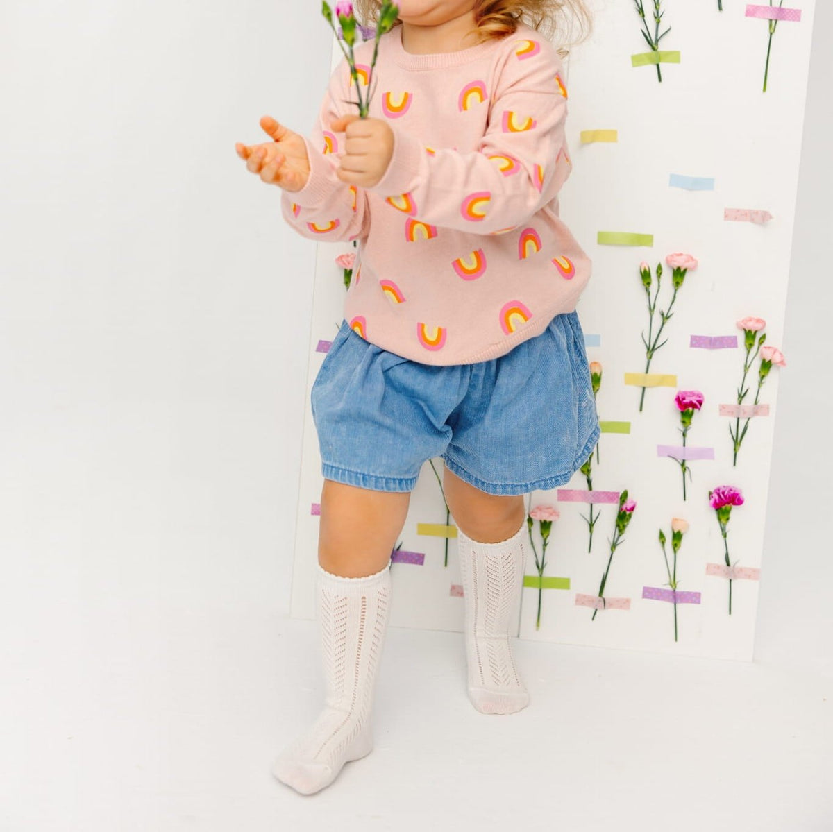 Kind met witte Toni kniekousen, een blauwe short en een roze trui met regenbogen, met bloemen in de hand.