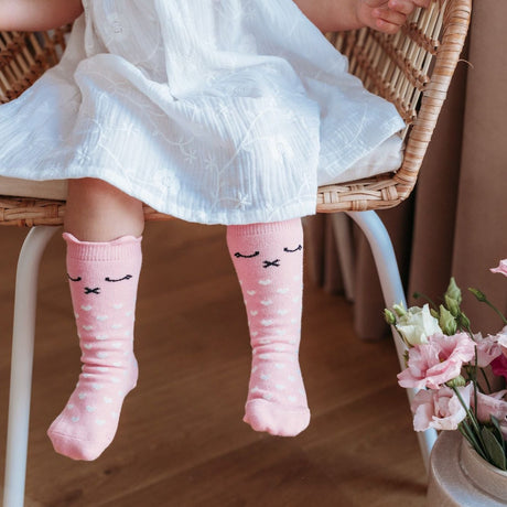 Roze Lilly a Pinky Kitty kniekousen met kattengezichtjes en witte hartjes, gedragen onder een witte jurk.