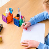 Kind tekent aan tafel naast twee pennenbakjes gemaakt van kleurrijke magnetische bouwblokken