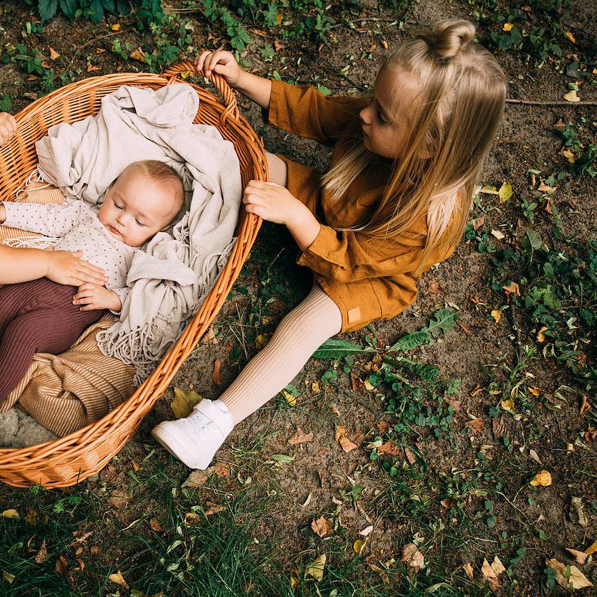 Kind in beige geribde maillot en okerkleurige jurk naast een rieten mand met een baby.