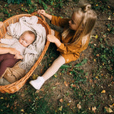 Kind in beige geribde maillot en okerkleurige jurk naast een rieten mand met een baby.