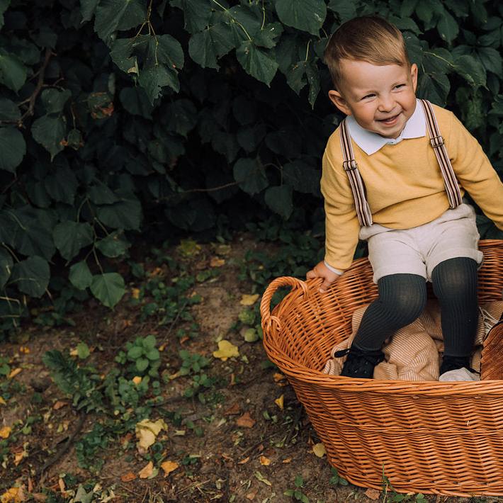 Kind met donkerkhaki geribde kniekousen, een gele trui en een korte broek, buiten zittend in een rieten mand.