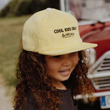 Kind met de gele Mini Citrus-pet van Hello Hossy met platte klep, bij een rode auto.