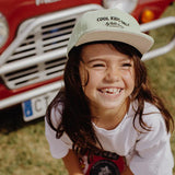 Lachend kind met de lichtgroene Mini Kiwi-pet van Hello Hossy, met een rode auto op de achtergrond.