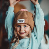 Lachend kind met bruine Block Macaroon muts met roze pompon en label vooraan, met de armen omhoog.
