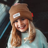 Lachend kind met bruine Block Macaroon muts van Hello Hossy, met omgeslagen rand en roze pompon.