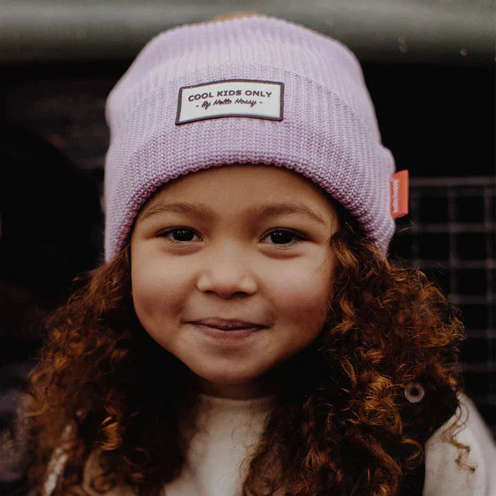 Kind met krullend haar draagt de lila Hello Hossy Block Mauve muts en glimlacht naar de camera.
