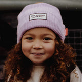 Kind met krullend haar draagt de lila Hello Hossy Block Mauve muts en glimlacht naar de camera.
