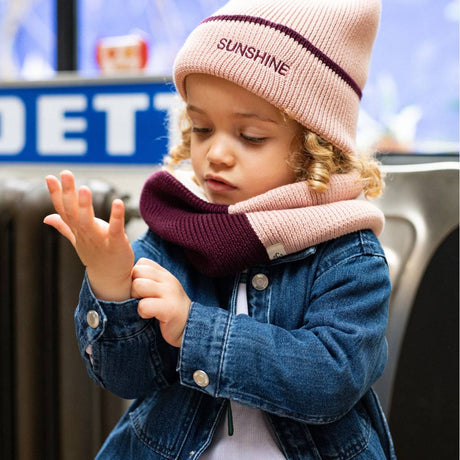 Kind met roze Sunshine-muts, roze en pruimkleurige sjaal en een spijkerjas.