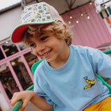 Kind op een groene stoel met de Hello Hossy Adventure pet, een lichtblauw shirt en een gele broek.
