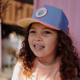 Glimlachend kind met blauwe Pet Patch Fleet van Hello Hossy, met ronde patch en roze klep met gele onderkant.