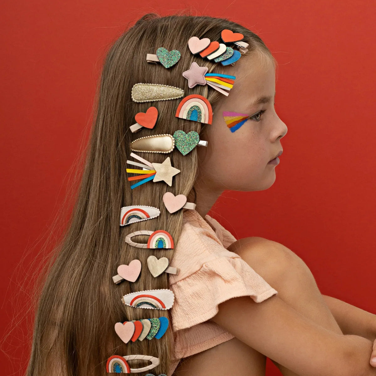 Meisje met lang haar vol haarclips met hartjes, sterren en regenbogen tegen een rode achtergrond.