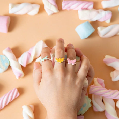 Drie goudkleurige ringen met een lichtgroene libel en gele en roze vlinders aan een hand, omringd door marshmallows.