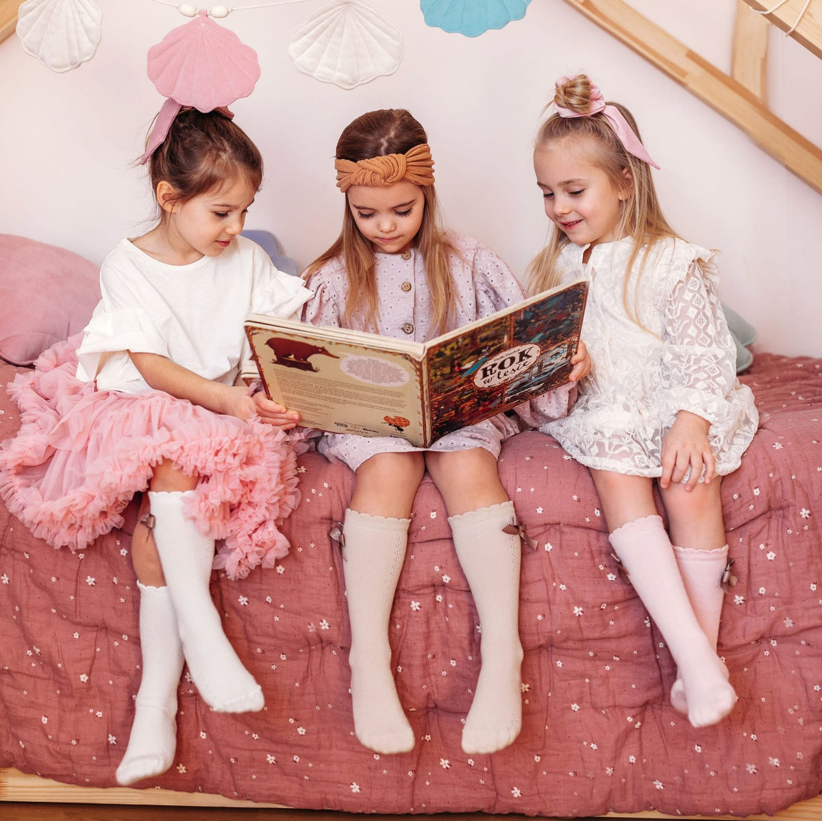 Drie meisjes met witte en lichtroze kniekousen met strikjes lezen samen een boek op een bed.
