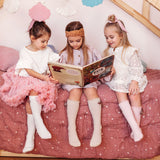 Drie meisjes met witte en lichtroze kniekousen met strikjes lezen samen een boek op een bed.