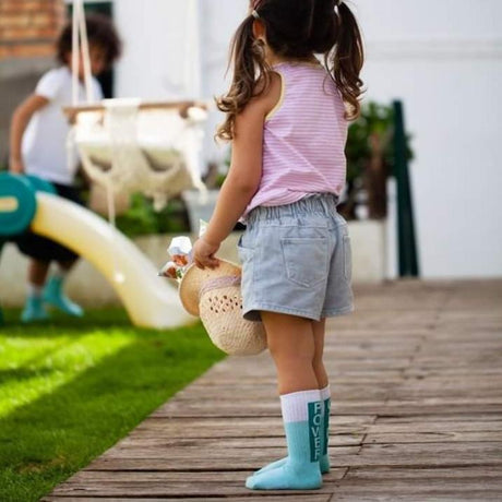 Kind met blauwe Girl Power sokken, een korte broek en een hoed in de hand op een houten terras.