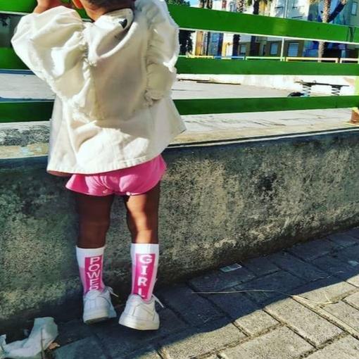 Kind met roze Girl Power-sokken en witte sneakers bij een stenen muur en groen hek.