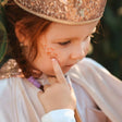 Kind met een glinsterende kroon wijst naar Star Dust Rainbow gezichtsglitter op de wang.