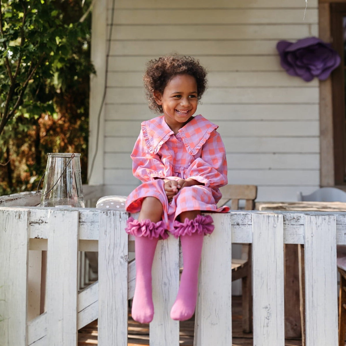 Kind met roze tutu kniekousen en een geruite jurk, zittend op een witte houten omheining.