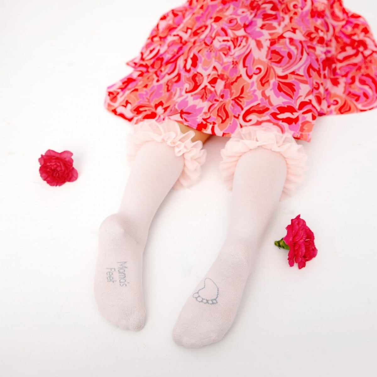 Kind draagt lichtroze Tutu kniekousen met ruches onder een roze-rode bloemenrok, met bloemen ernaast.