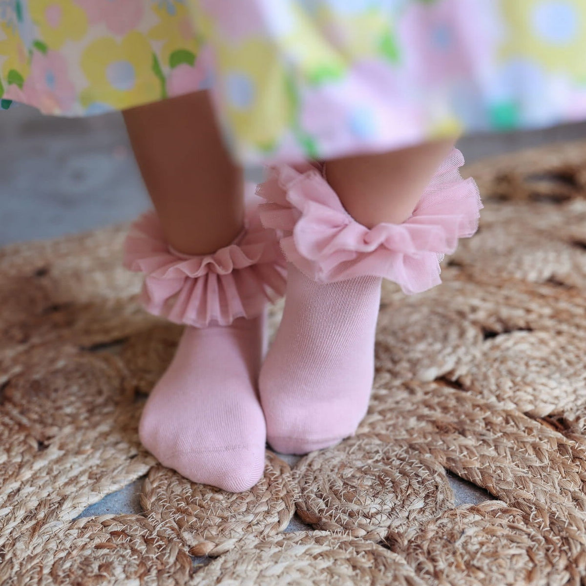 Oudroze tutu sokken met ruches, gedragen onder een gebloemde jurk op een gevlochten kleed.