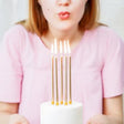 Vrouw blaast naar brandende gouden verjaardagskaarsen op een witte taart.