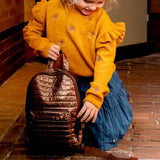Kind met gele trui houdt de glanzende Walker rugtas in Dark Gold vast bij de draaglus.