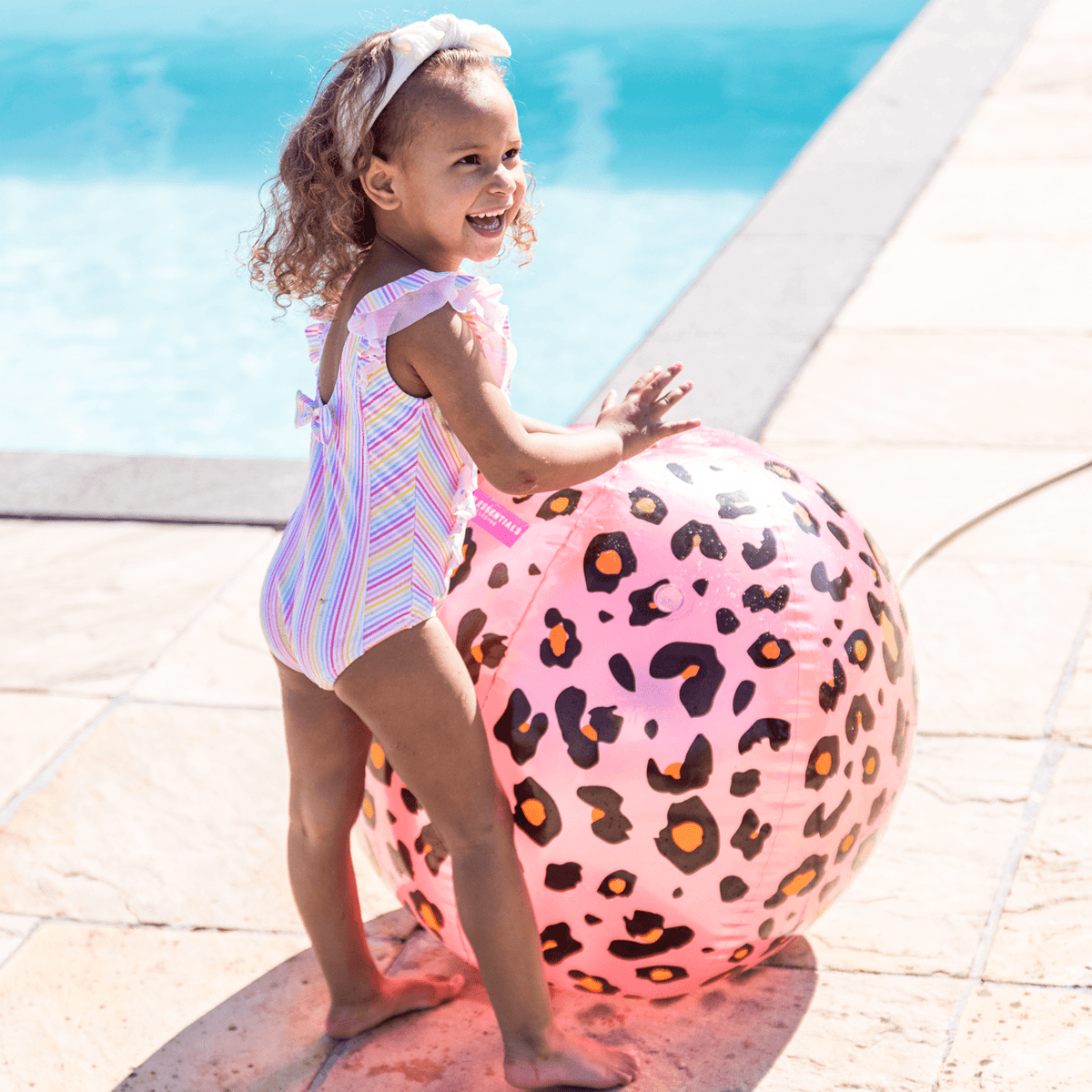 Kind met een hand op de roze, bolvormige watersproeier met panterprint naast het zwembad.