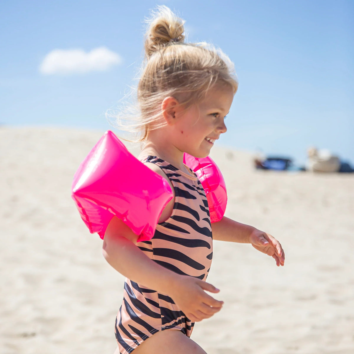 Kind met neonroze zwembandjes en een badpak met dierenprint op het strand, van opzij gezien.