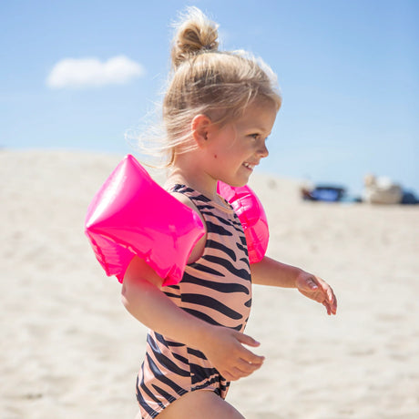 Kind met neonroze zwembandjes en een badpak met dierenprint op het strand, van opzij gezien.