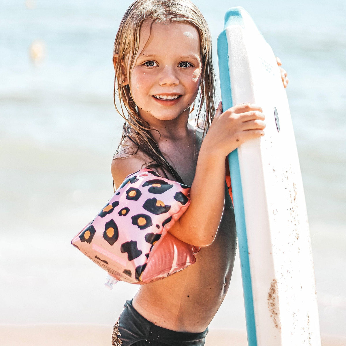 Meisje met roze zwembandjes met panterprint houdt een wit met blauw board vast op het strand.